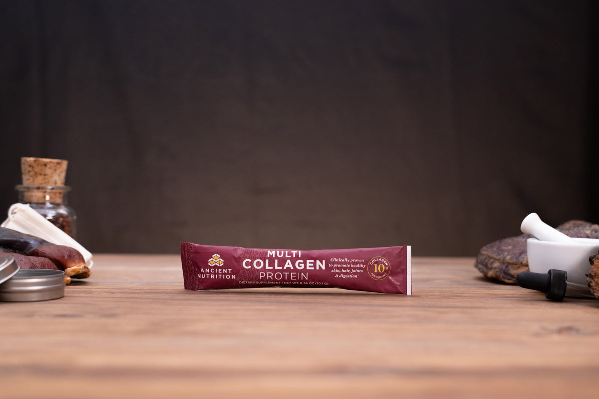 A tube of Ancient Nutrition Multi Collagen Protein powder lies on a wooden tabletop, with blurred natural props including a cork bottle, dried herbs, and a white mortar in the background.
