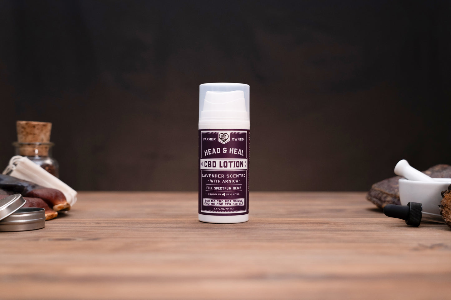 A bottle of Head & Heal Red Lotion stands centered on a wooden tabletop against a dark background, with small cosmetic and natural props visible on both sides.