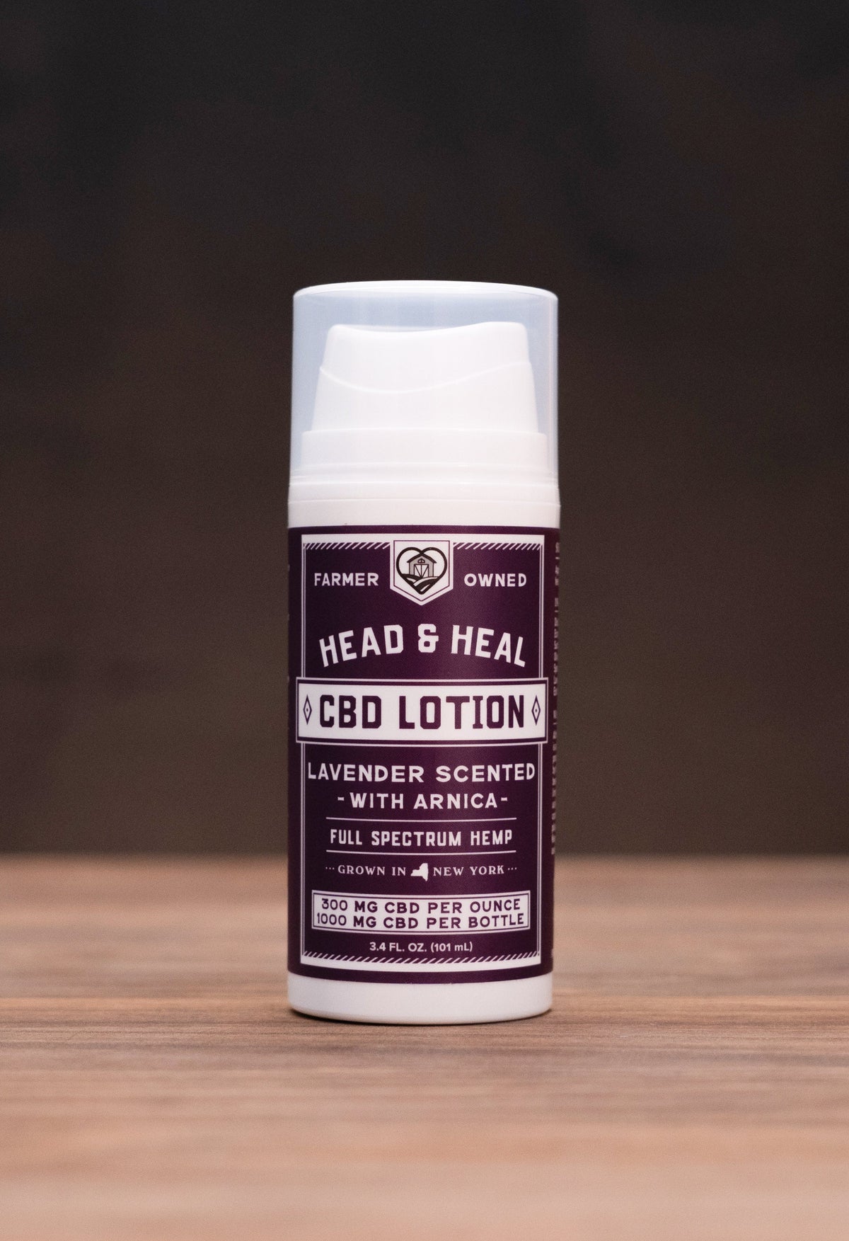 A bottle of Head & Heal Red Lotion stands centered on a wooden tabletop against a dark background, with small cosmetic and natural props visible on both sides.
