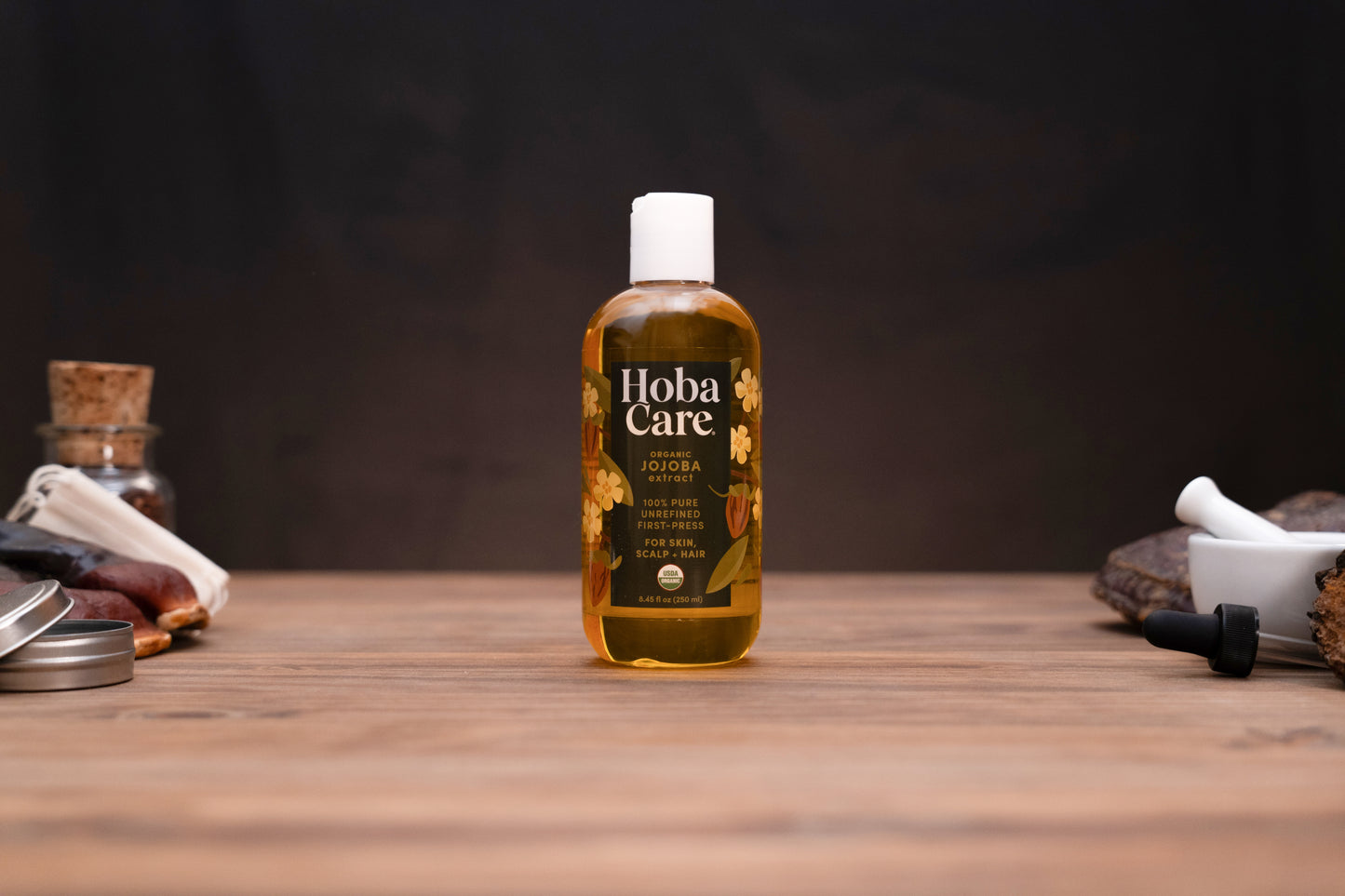 A bottle of Hoba Care Sacred Jojoba Oil stands centered on a rustic wooden table, surrounded by traditional herbal props against a dark, moody background.