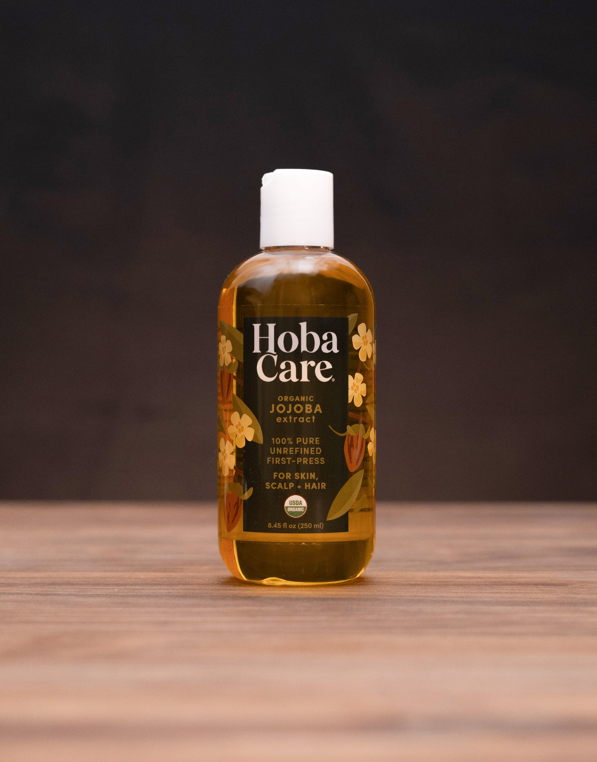 A bottle of Hoba Care Sacred Jojoba Oil stands centered on a rustic wooden table, surrounded by traditional herbal props against a dark, moody background.