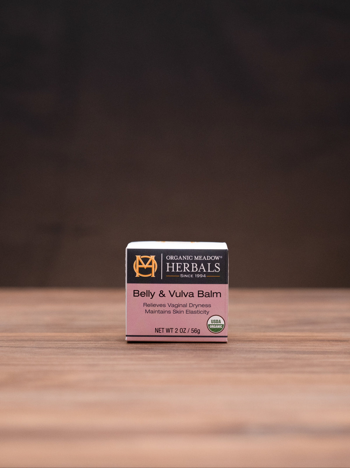 A small boxed product labeled 'The Herbalist Baby & Vena Barn' sits centered on a wooden tabletop against a dark brown backdrop, with blurred herbal props on both sides.