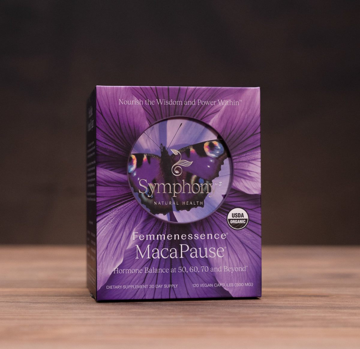 Symphonics Maca Pause supplement box in purple packaging, standing on a wooden surface with blurred herbal props in the background.