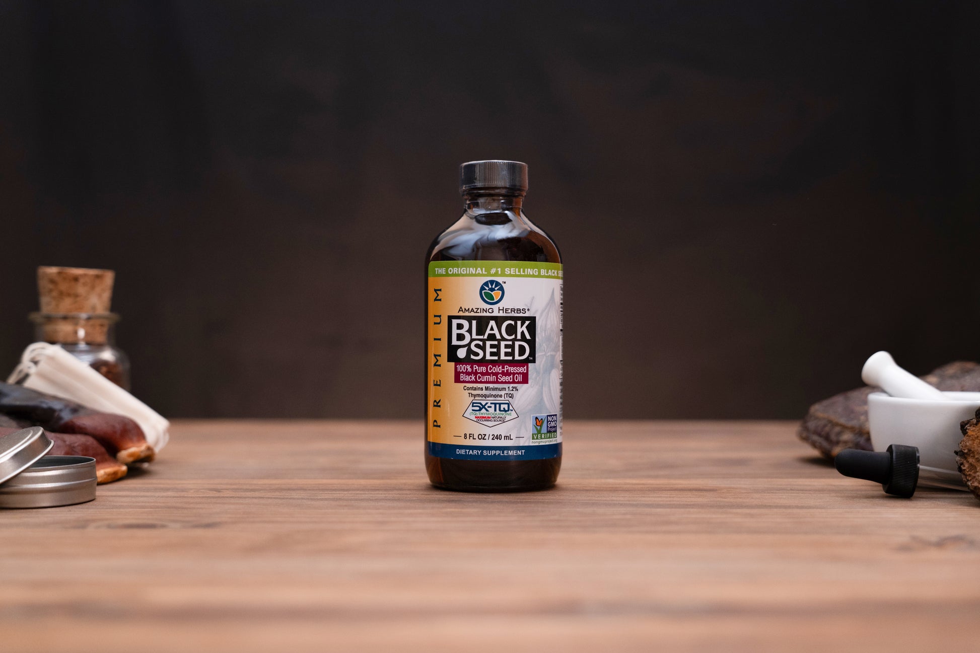 A bottle of K2 Health Black Seed supplement standing on a wooden tabletop, with small herbal props blurred in the background against a dark backdrop.