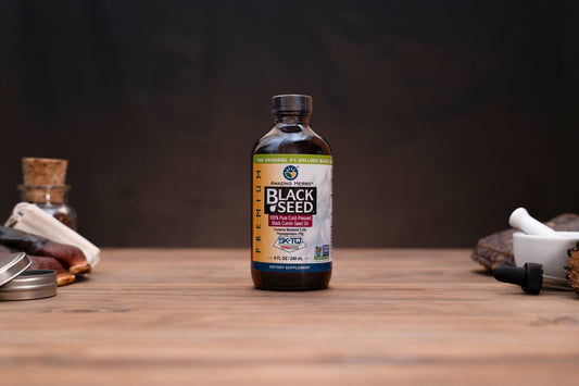 A bottle of K2 Health Black Seed supplement standing on a wooden tabletop, with small herbal props blurred in the background against a dark backdrop.