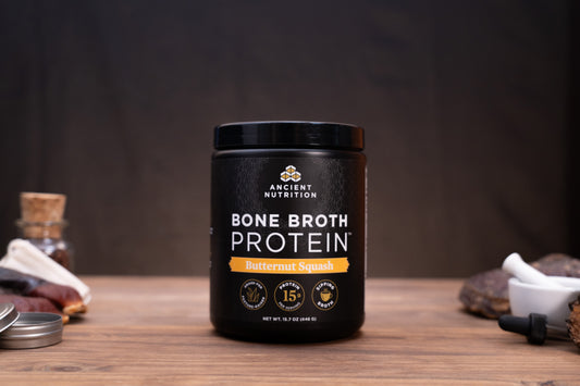 A black tub of Ancient Nutrition Bone Broth Protein in Butternut Squash flavor sits on a wooden table with rustic ingredients and a dark background.