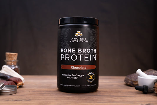 A black tub of Ancient Nutrition Bone Broth Protein in Chocolate flavor, displayed on a wooden surface with rustic ingredients and accessories in the background.