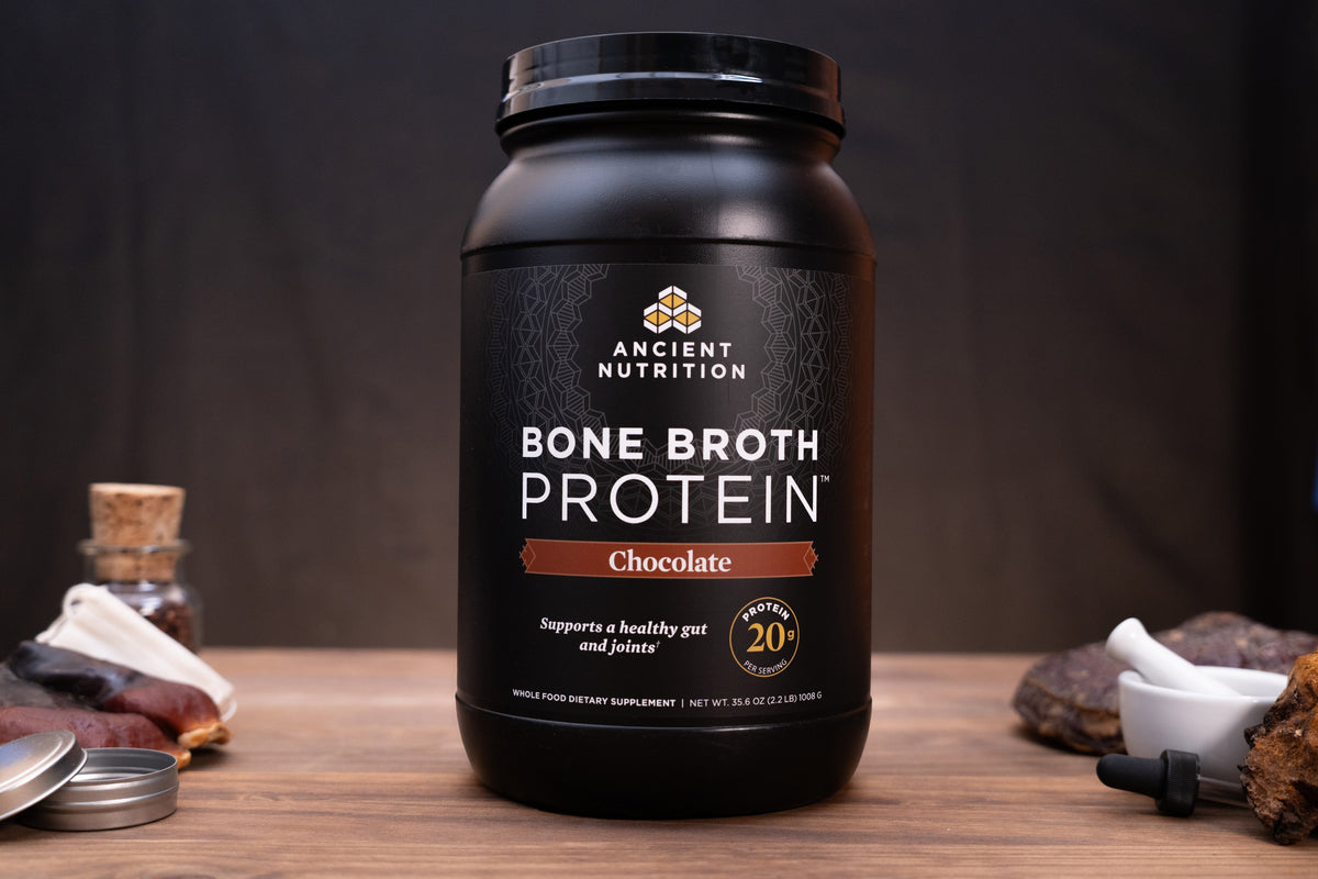 A black tub of Ancient Nutrition Bone Broth Protein in Chocolate flavor sits on a wooden surface with rustic ingredients and a small white mortar in the background.