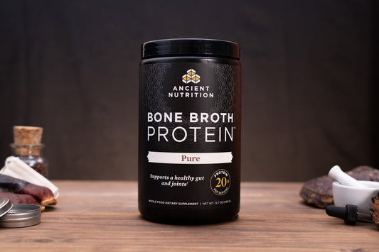 A black plastic tub of Ancient Nutrition Bone Broth Protein Pure, set on a rustic wooden table with herbal props in the background.