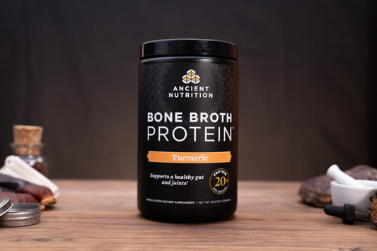 A black plastic tub of Ancient Nutrition Bone Broth Protein in Turmeric flavor, set on a rustic wooden table with various herbal and kitchen props blurred in the background.