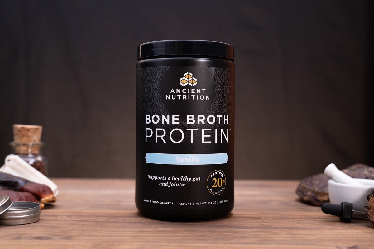 A black tub of Ancient Nutrition Bone Broth Protein Vanilla flavor sits on a wooden table with rustic props like a small glass bottle, dried herbs, and a mortar and pestle against a dark background.