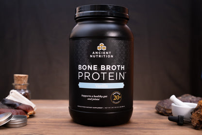 A black tub of Ancient Nutrition Bone Broth Protein in Vanilla flavor sits on a wooden table with small food props like dried fruit, a small bottle, and a dropper around it against a dark background.