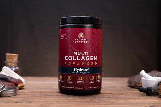 A bottle of Ancient Nutrition Multi Collagen Advanced dietary supplement, set on a rustic wooden table with natural elements like a cork stopper, dried herbs, and a dropper in the background.