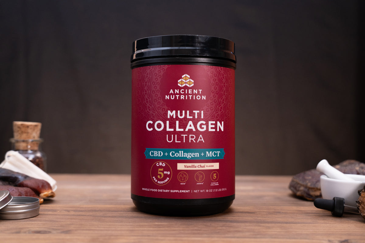 A black plastic tub of Ancient Nutrition Multi Collagen Ultra Vanilla Chai Spice on a wooden table, surrounded by small rustic props like a cork stopper, a small jar, and a white mortar and pestle against a dark background.