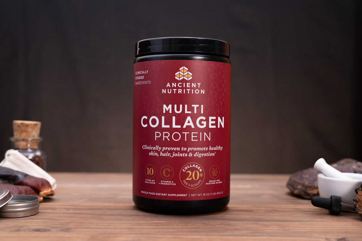 A black plastic tub of Ancient Nutrition Multi Collagen Protein powder with a deep red label, displayed on a wooden surface with herbal and supplement props in the background.