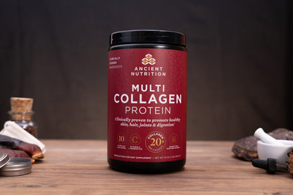 A black plastic tub of Ancient Nutrition Multi Collagen Protein powder with a deep red label, displayed on a wooden surface with herbal and supplement props in the background.