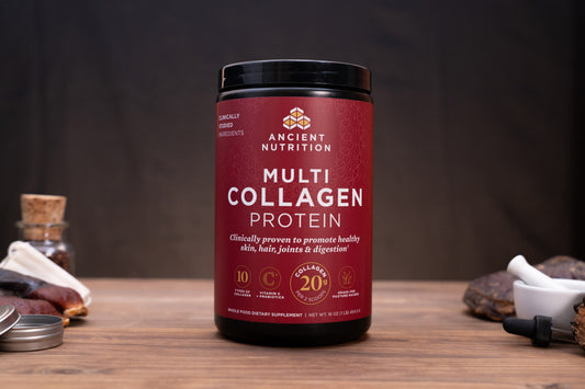 A black plastic tub of Ancient Nutrition Multi Collagen Protein powder with a deep red label, displayed on a wooden surface with herbal and supplement props in the background.