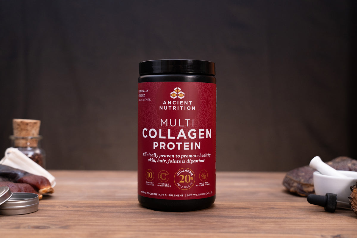 A jar of Ancient Nutrition Multi Collagen Protein powder sits on a wooden table with rustic props like a cork stopper, dried herbs, and a small white bowl against a dark background.