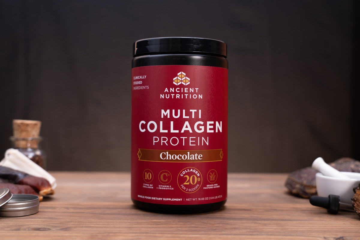 A jar of Ancient Nutrition Multi Collagen Protein powder in Chocolate flavor, displayed on a wooden surface with rustic ingredients in the background.