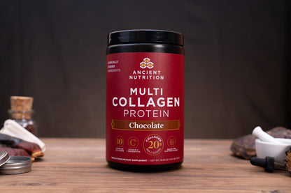 A jar of Ancient Nutrition Multi Collagen Protein powder in Chocolate flavor, displayed on a wooden surface with rustic ingredients in the background.