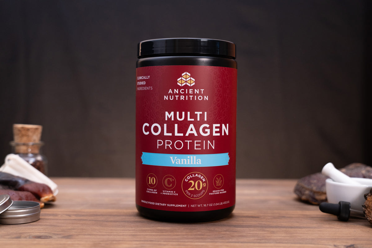 A jar of Ancient Nutrition Multi Collagen Protein powder in Vanilla flavor, displayed on a rustic wooden table with small herbal and supplement props in the background.