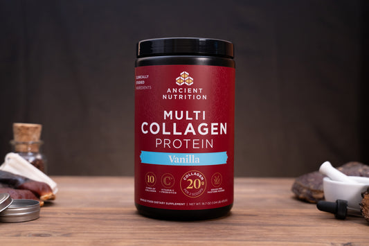 A jar of Ancient Nutrition Multi Collagen Protein powder in Vanilla flavor, displayed on a rustic wooden table with small herbal and supplement props in the background.