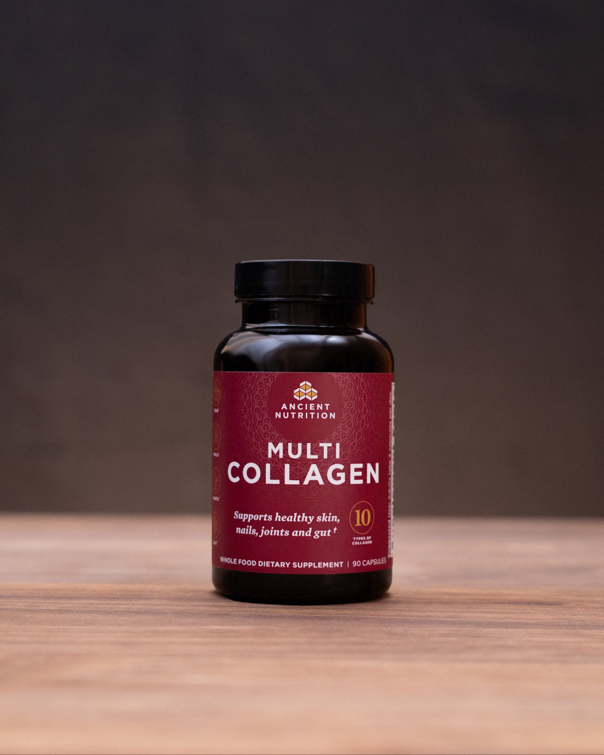 A bottle of Western Health Multi Collagen dietary supplement standing on a wooden surface, with blurred natural props on both sides against a dark background.