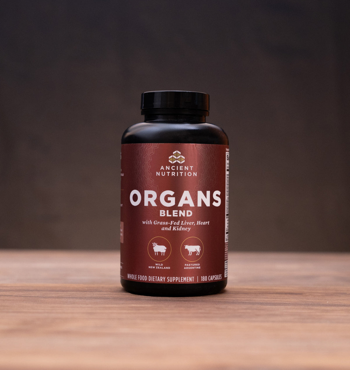 A bottle of Ancient Nutrition Organs Blend supplement stands on a wooden table against a dark background, surrounded by small rustic props including a cork stopper, metal lid, and white mortar and pestle.