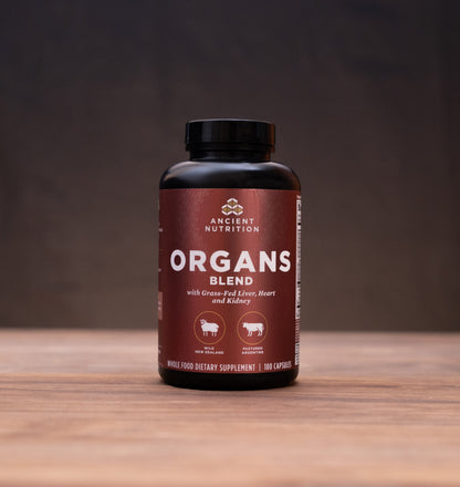 A bottle of Ancient Nutrition Organs Blend supplement stands on a wooden table against a dark background, surrounded by small rustic props including a cork stopper, metal lid, and white mortar and pestle.