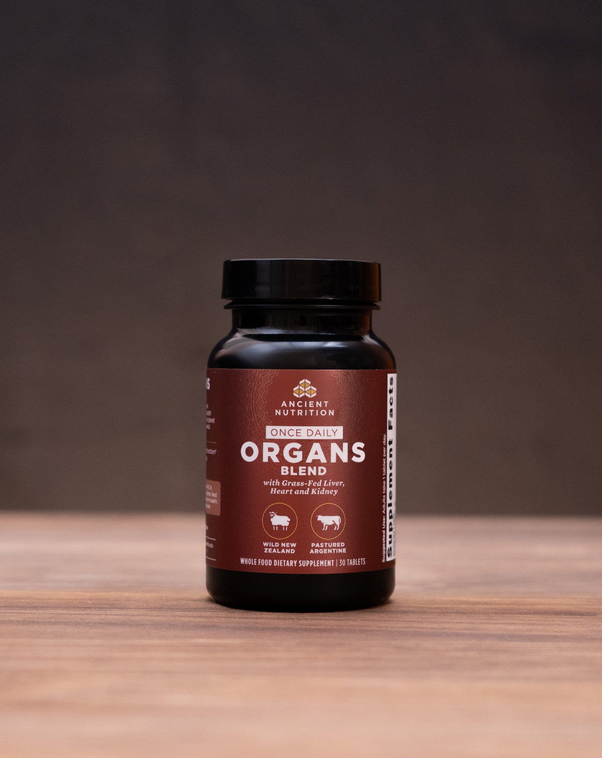 A dark amber bottle of Organ's Organics supplement stands centered on a wooden tabletop, surrounded by blurred natural and kitchen props including a cork stopper, dried herbs, and a white mortar and pestle against a dark background.