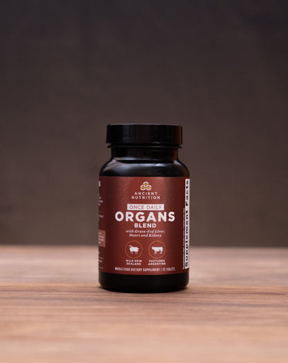 A dark amber bottle of Organ's Organics supplement stands centered on a wooden tabletop, surrounded by blurred natural and kitchen props including a cork stopper, dried herbs, and a white mortar and pestle against a dark background.
