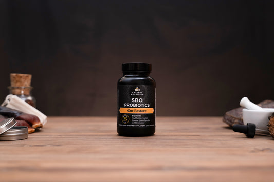 A black bottle of SBO Probiotics Gut Restore dietary supplement centered on a wooden tabletop, with blurred herbal props on both sides against a dark background.