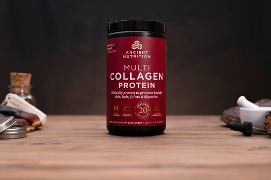 A bottle of Ancient Nutrition Multi Collagen Protein powder stands on a wooden surface against a dark background, with small blurred props on both sides.