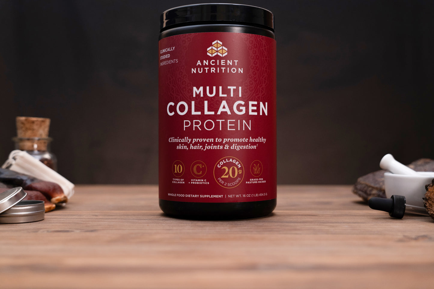 A bottle of Ancient Nutrition Multi Collagen Protein powder on a wooden table with a dark background and small props.