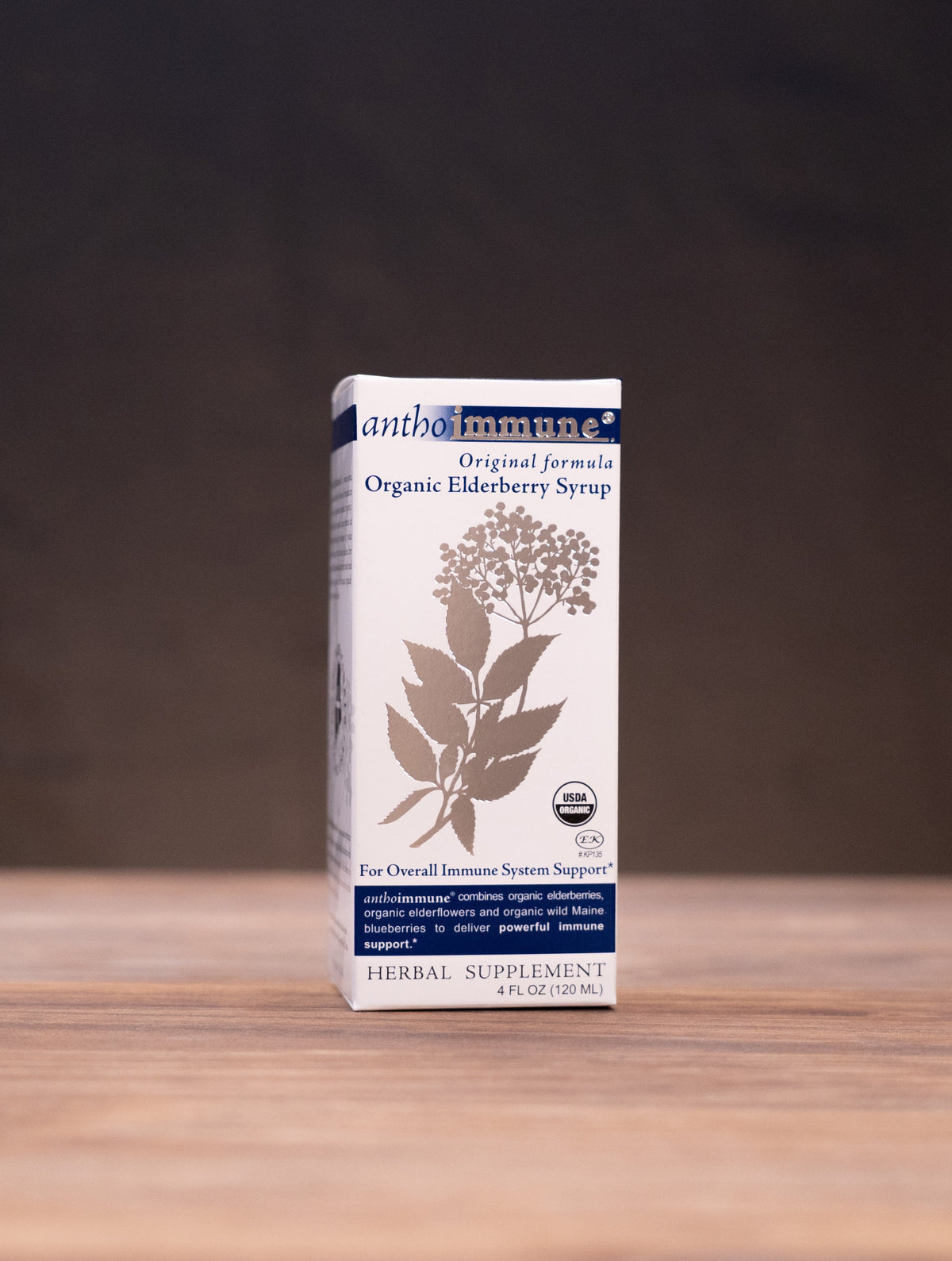 A box of andalamar Organic Elderberry Syrup stands centered on a wooden tabletop against a dark backdrop, with blurred jars and a mortar-and-pestle props visible on both sides.