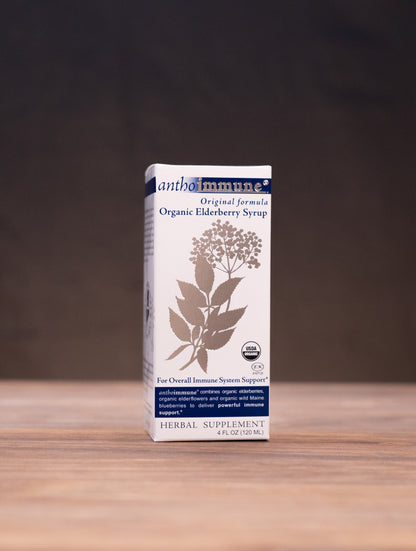 A box of andalamar Organic Elderberry Syrup stands centered on a wooden tabletop against a dark backdrop, with blurred jars and a mortar-and-pestle props visible on both sides.
