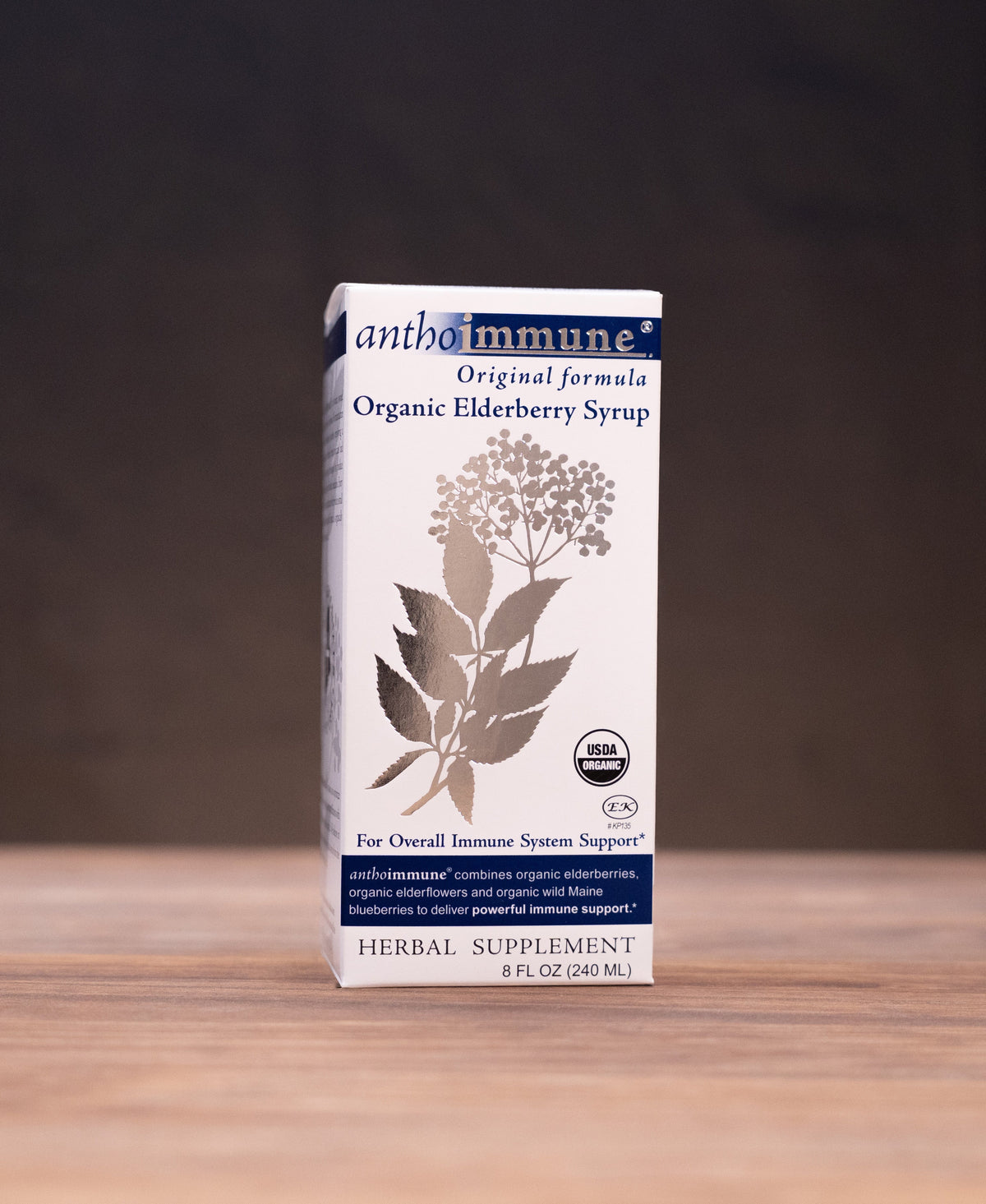 A box of Ambimuni Organic Elderberry Syrup stands on a wooden tabletop against a dark backdrop, with small herbal-themed props visible on both sides.