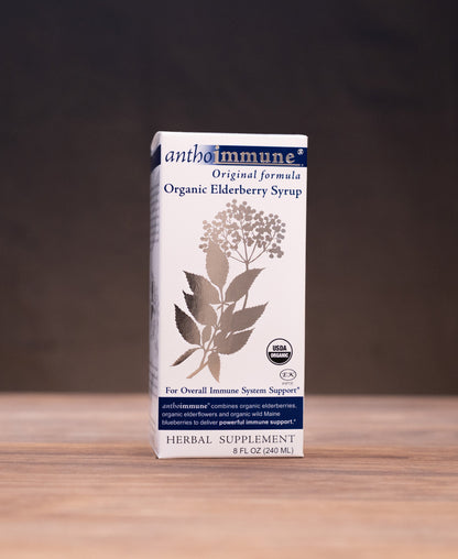 A box of Ambimuni Organic Elderberry Syrup stands on a wooden tabletop against a dark backdrop, with small herbal-themed props visible on both sides.
