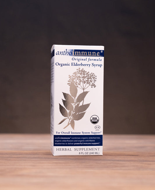 A box of Ambimuni Organic Elderberry Syrup stands on a wooden tabletop against a dark backdrop, with small herbal-themed props visible on both sides.