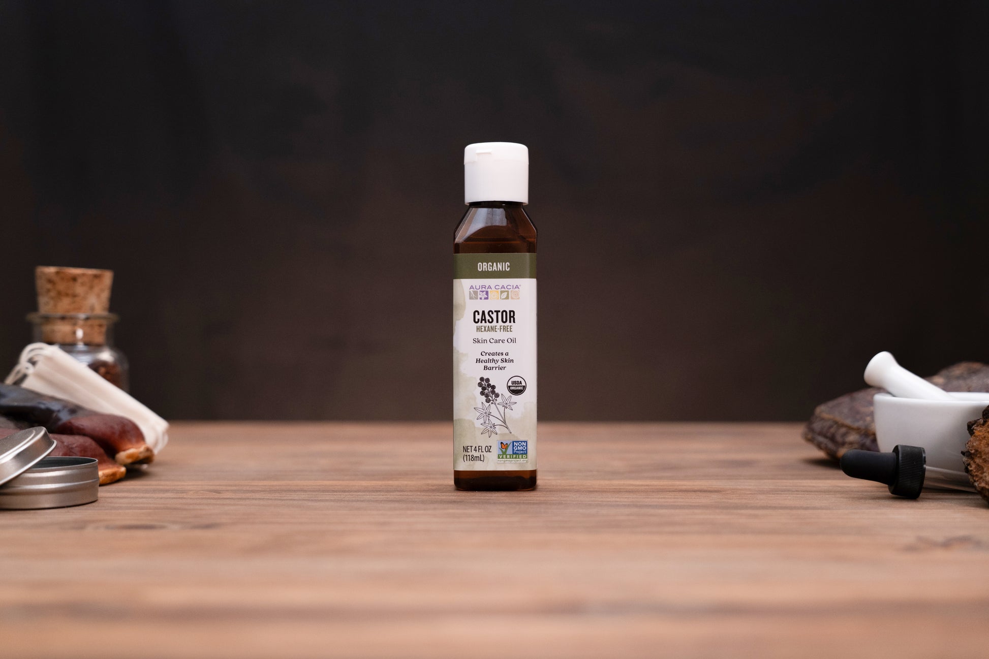 A small amber bottle of Castor oil stands centered on a wooden tabletop against a dark background, with blurred herbal and laboratory-style props visible on both sides.