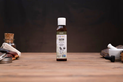 A small amber bottle of Castor oil stands centered on a wooden tabletop against a dark background, with blurred herbal and laboratory-style props visible on both sides.