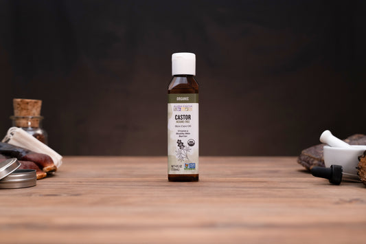 A small amber bottle of Castor oil stands centered on a wooden tabletop against a dark background, with blurred herbal and laboratory-style props visible on both sides.