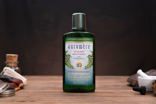 A bottle of auromère Antiviral Mouthwash with Neem & Pelti, set on a wooden surface with herbal props in the background.