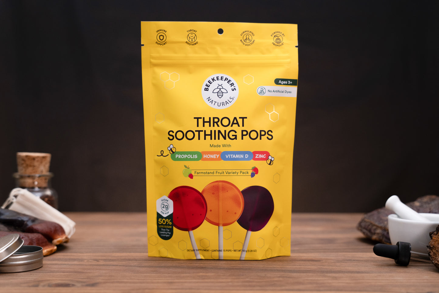 A yellow stand-up pouch of Beekeeper throat-soothing pops candy stands on a wooden surface against a dark background. The package shows three illustrated lollipops in red, orange, and dark purple, with text including 'Throat Soothing Pops' and 'Ages 3+'.