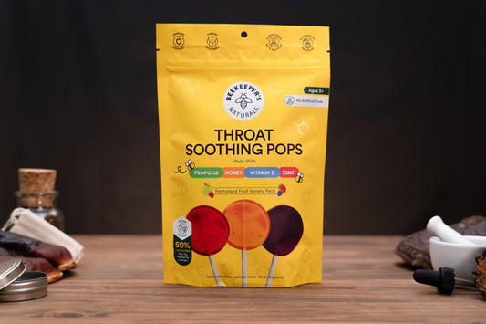 A yellow stand-up pouch of Beekeeper throat-soothing pops candy stands on a wooden surface against a dark background. The package shows three illustrated lollipops in red, orange, and dark purple, with text including 'Throat Soothing Pops' and 'Ages 3+'.