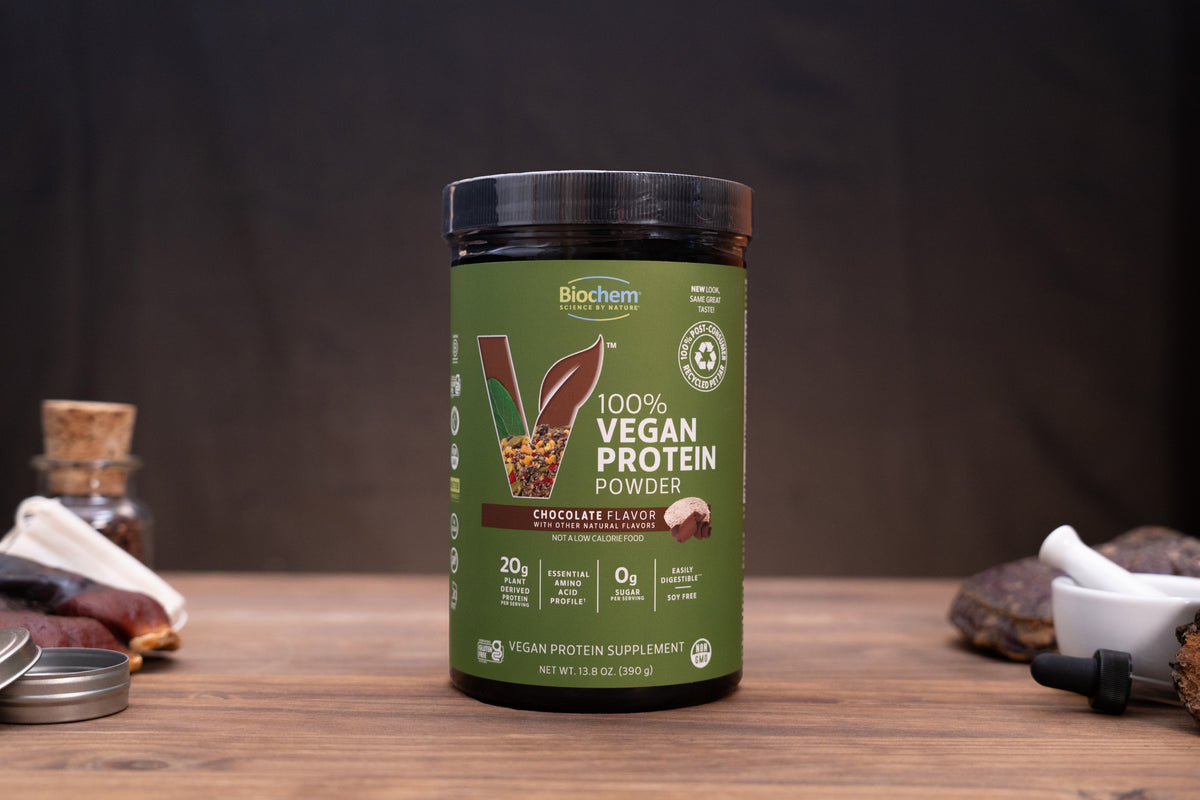 Biochem 100% Vegan Protein Powder in Chocolate Flavor, in a dark green tub with a black lid, displayed on a wooden surface with small ingredient props on either side against a dark background.