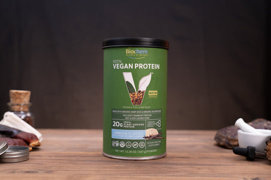 A canister of Biochem 100% Vegan Protein in Vanilla flavor, set on a wooden table with rustic ingredients like cocoa, a mortar and pestle, and a small bottle in the background.