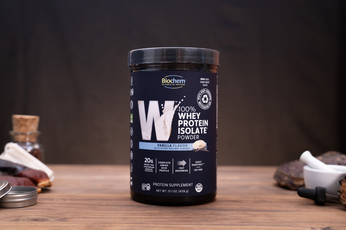 Biochems Vanilla Flavor Whey Protein Isolate powder in a black tub, displayed on a wooden surface with small kitchen props in the background.
