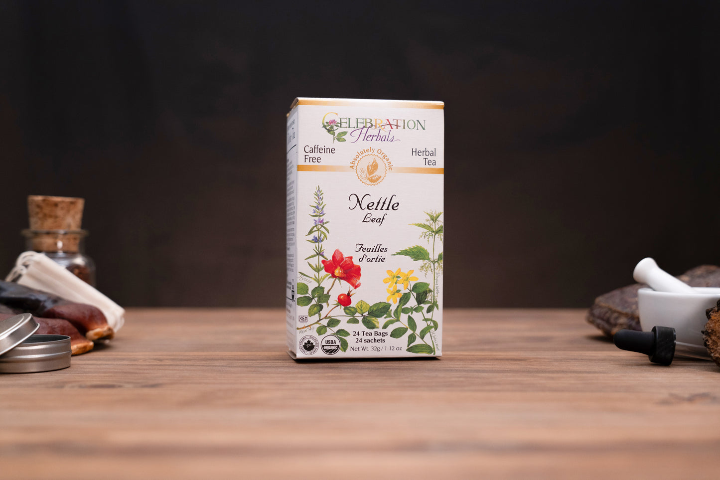 A box of Celebration Herbs Kettle Leaf herbal tea stands centered on a wooden tabletop against a dark background, with small blurred props on both sides.