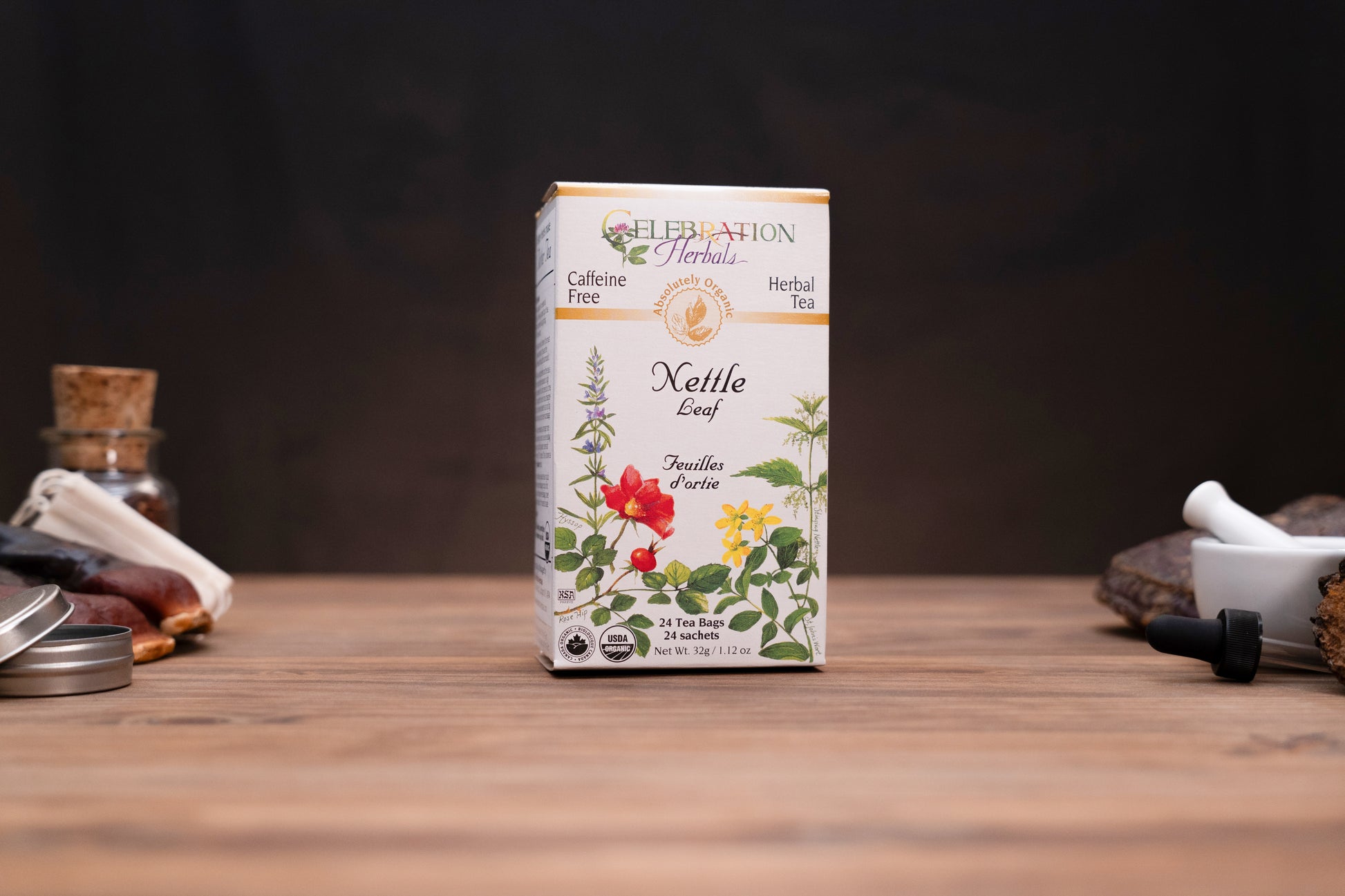 A box of Celebration Herbs Kettle Leaf herbal tea stands centered on a wooden tabletop against a dark background, with small blurred props on both sides.